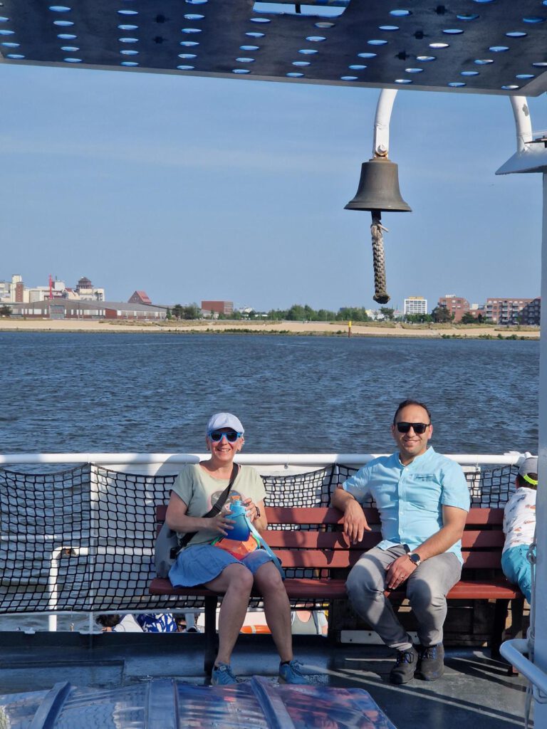 Zwei Personen mit Sonnenbrillen sitzen auf einer Bank auf einer Fähre. Im Hintergrund sind Wasser und ein Sandstrand zu sehen.