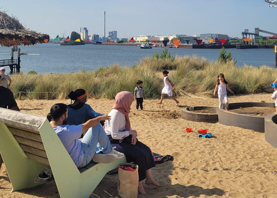 An einem Sandstrand sind Bänke mit Erwachsenen und Kindern zu sehen. Im Hintergrund gibt es Wasser.