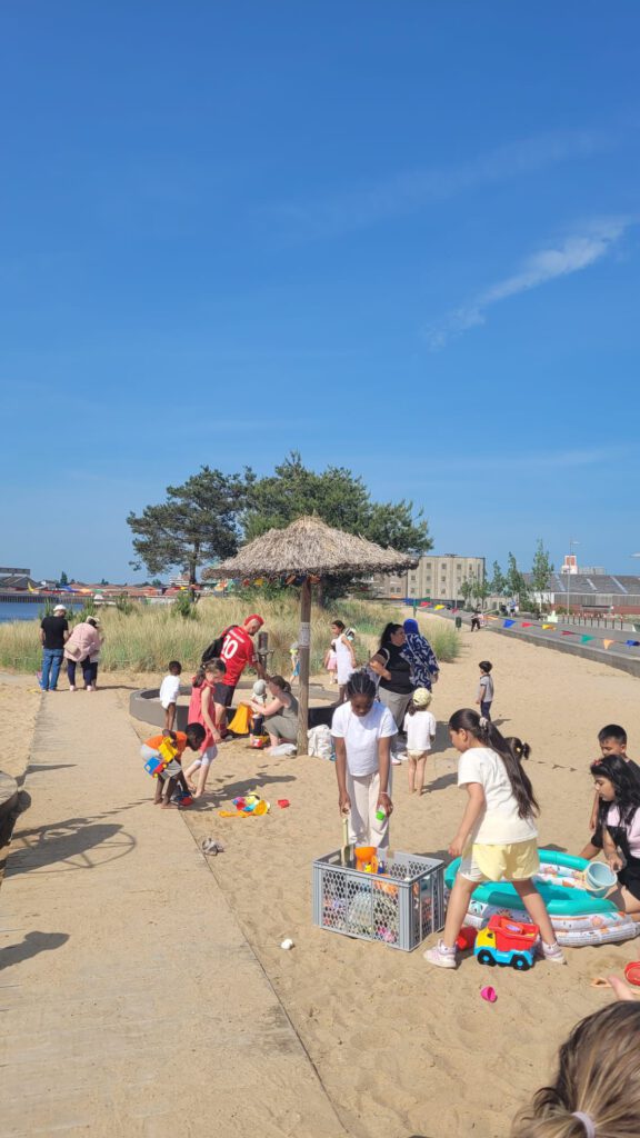 An einem Sandstrand sind Bänke mit Erwachsenen und Kindern zu sehen. Im Hintergrund gibt es Wasser.
