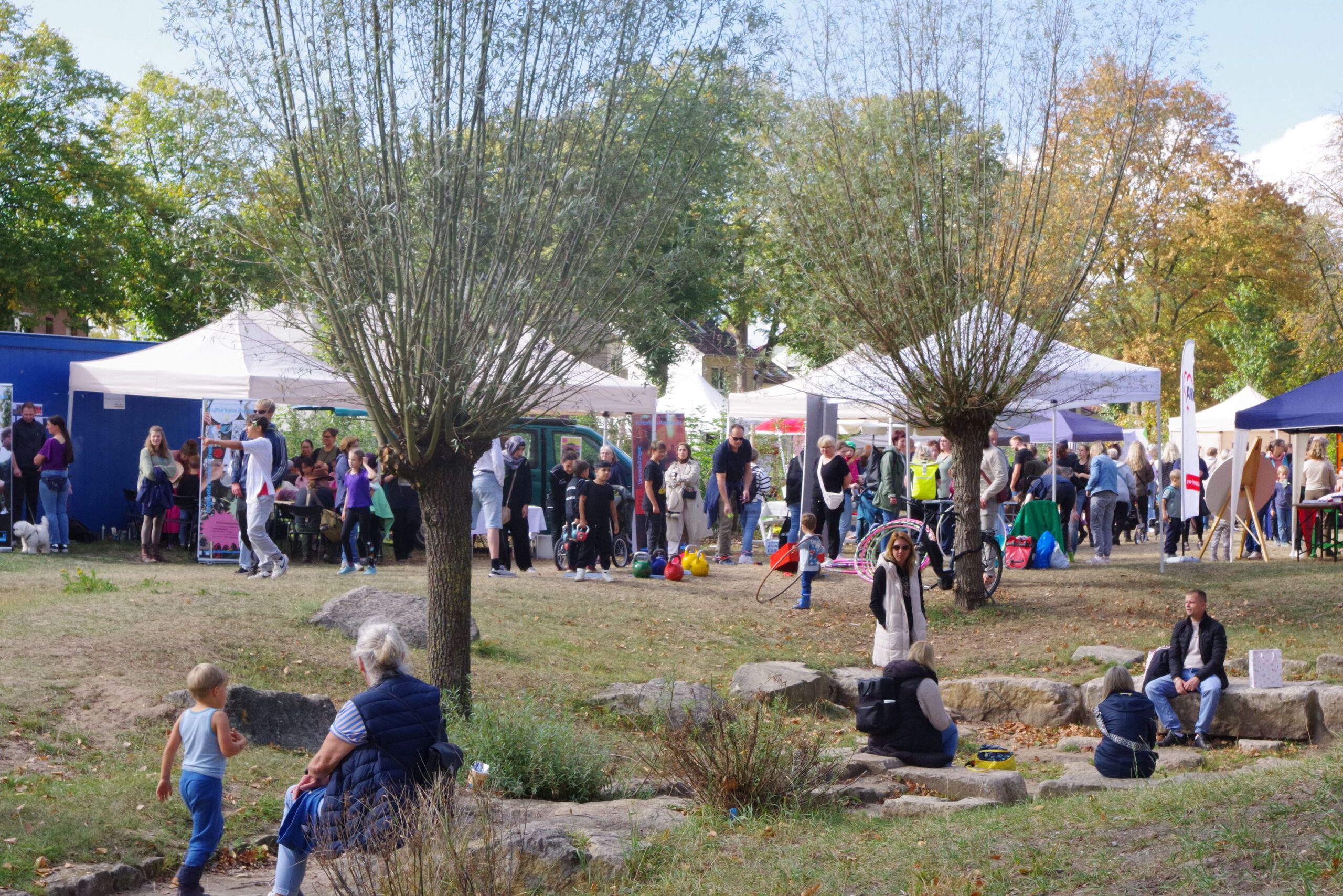 Auf einer großen wiese mit Weiden findet ein Stadtteilfest mit vielen Pavillonen statt. Menschen sitzen auf Piknickdecken