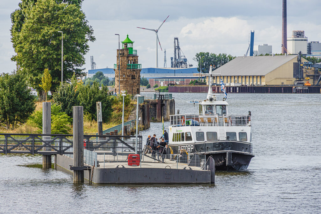 Ein weiß‑schwarzes Fahrgastschiff mit dem Namen „PUSDORP“ liegt an einem Steg. Mehrere Personen stehen auf dem Boot oder auf einem Schwimmanleger. Im Hintergrund stehen ein Leuchtturm mit grünem Dach, Bäume sowie Industrieanlagen mit Kränen und einer Windkraftanlage. Das Wasser ist ruhig, der Himmel teils bewölkt.