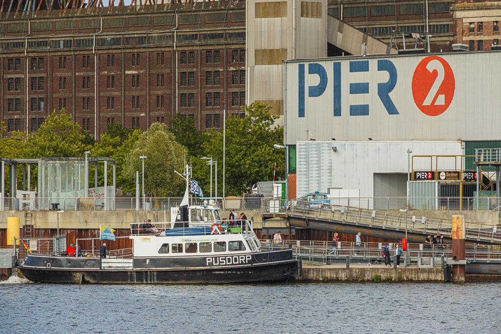 Am Ufer liegt ein schwarz‑weißes Schiff mit dem Namen „PUSDORP“ an einem Gebäude mit der großen Aufschrift „PIER 2“. Auf dem Steg und in der Nähe des Eingangsbereichs bewegen sich mehrere Personen. Hinter dem Pier steht ein großes Industriegebäude mit vielen Fenstern. Bäume und weitere Hafenstrukturen rahmen die Szene, der Himmel ist bewölkt.
