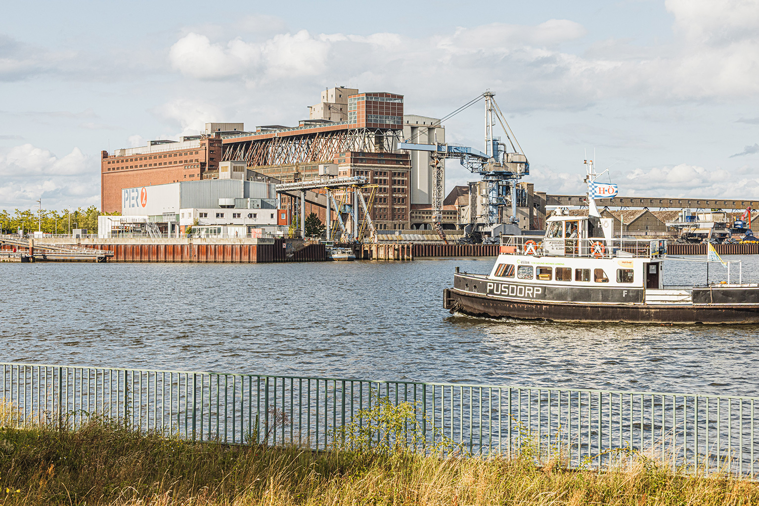 Eine schwarz‑weiße Fähre mit dem Namen „PUSDORP“ fährt durch ein Hafenbecken. Im Hintergrund stehen große Industriegebäude, Silos und metallene Hafenanlagen, darunter ein Gebäude mit der Aufschrift „PIER 2“. Im Vordergrund verläuft ein grasbewachsener Uferstreifen mit einem Metallzaun. Der Himmel ist dicht bewölkt.