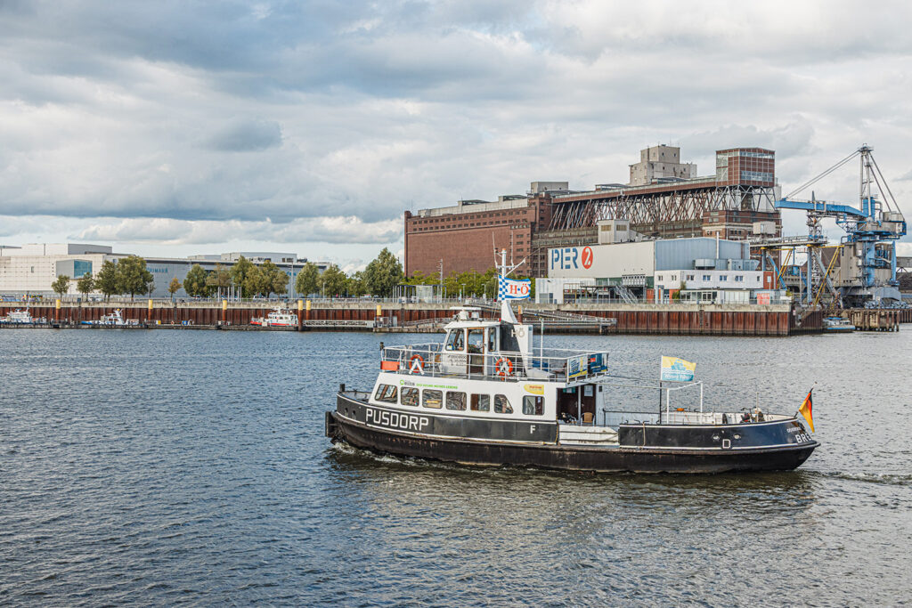 Auf einem Gewässer fährt eine kleine Personenfähre mit dem Namen „PUSDORP“. Das Schiff ist schwarz‑weiß lackiert und trägt mehrere Flaggen, darunter eine am Heck. Im Hintergrund stehen Industriegebäude, Kräne und ein großes Gebäude mit der Aufschrift „PIER 2“ unter einem teils bewölkten Himmel.