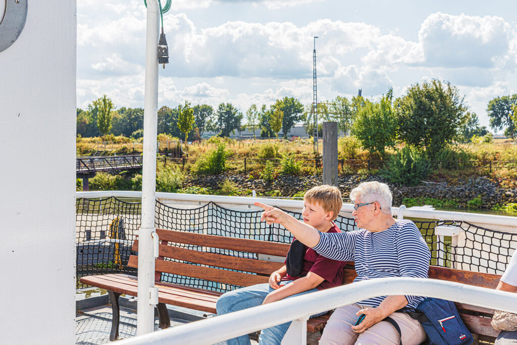 Auf einem Fährdeck sitzen eine ältere und eine junge Person auf einer hölzernen Bank und blicken in dieselbe Richtung. Die ältere Person hebt einen Arm und zeigt auf einen Punkt in der Ferne, während ein Smartphone in der anderen Hand gehalten wird. Hinter ihnen verläuft ein Uferstreifen mit Gras, Bäumen, einer kleinen Brücke und einem hohen Mast. Der Himmel ist hell und teils bewölkt.