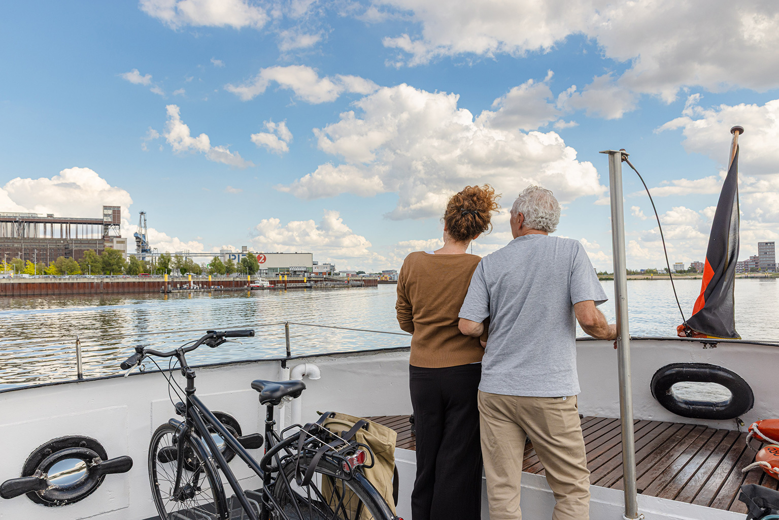 Ein Paar ist von hinten zu sehen. Sie stehen an Bord eines Schiffes, links von ihnen befindet sich ein Fahrrad mit Satteltasche. Sie blicken Richtung Ufer, bei dem ein großes Industriegebäude zu sehen ist und ein kleineres, auf dem "Pier 2" geschrieben steht. Am blauen Himmel sind Wolken.