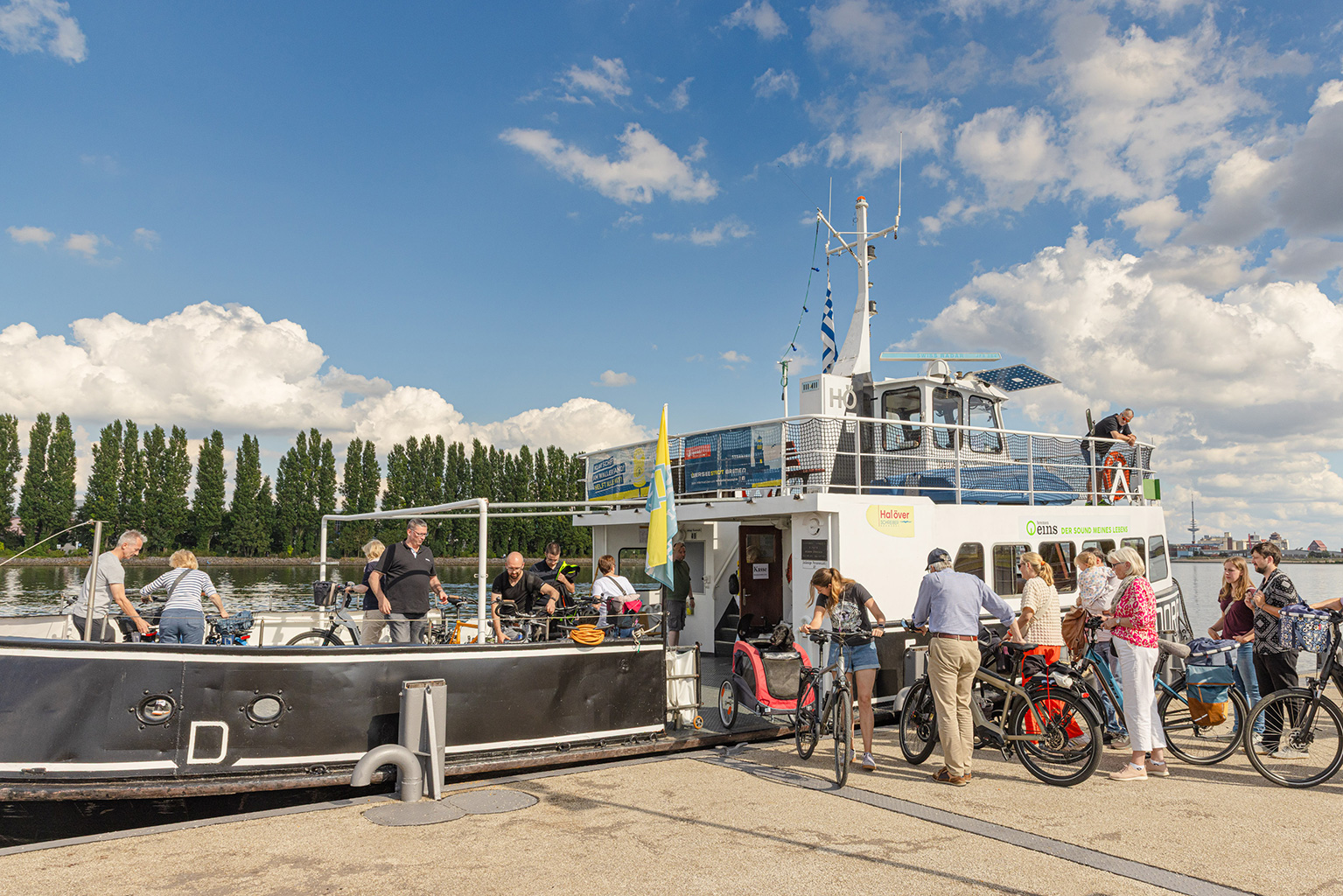An einem Anleger steigen Fußgänger und Radfahrende an Bord einer Fähre. Auf dem Schiff befinden sich schon viele Personen. Im Hintergrund ist eine Reihe Pappeln zu sehen.