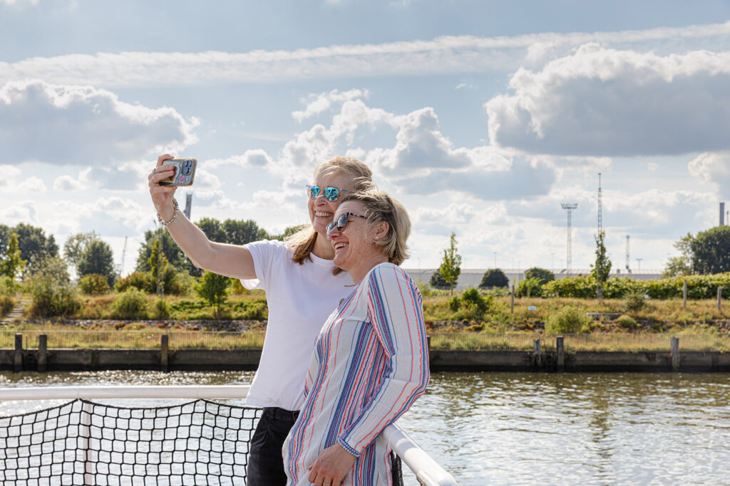 Zwei Personen stehen auf einem Boot oder Steg und halten ein Smartphone für ein Selfie in die Höhe. Eine Person trägt ein weißes T‑Shirt, die andere ein langärmliges Oberteil mit roten, blauen und weißen Streifen. Hinter ihnen liegt ein Uferbereich mit Bäumen, Grünflächen und einzelnen Industrieanlagen. Der Himmel ist teils bewölkt mit großen, hellen Wolkenformationen.