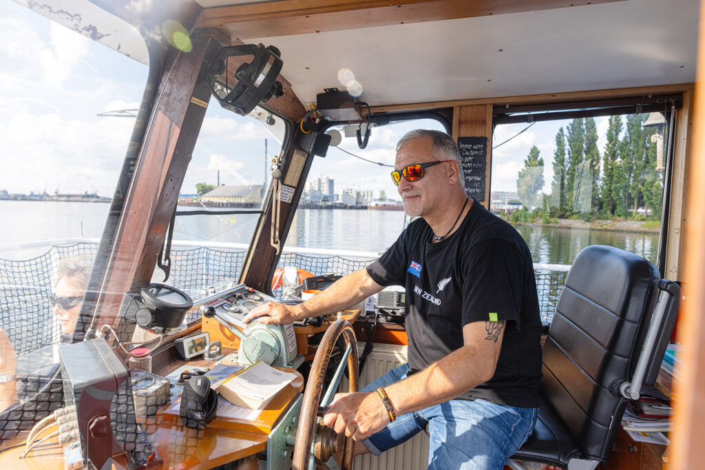In einem Steuerstand sitzt eine Person am Steuerrad eines Bootes und bedient gleichzeitig einen Hebel. Vor der Person liegen verschiedene Navigations- und Bedieninstrumente sowie ein aufgeschlagenes Buch und einige Papiere. Durch die Fenster ist ein ruhiges Gewässer mit einer Uferlinie aus Gebäuden und Bäumen zu sehen. An der Wand hängt eine kleine Tafel mit einem handgeschriebenen Spruch.