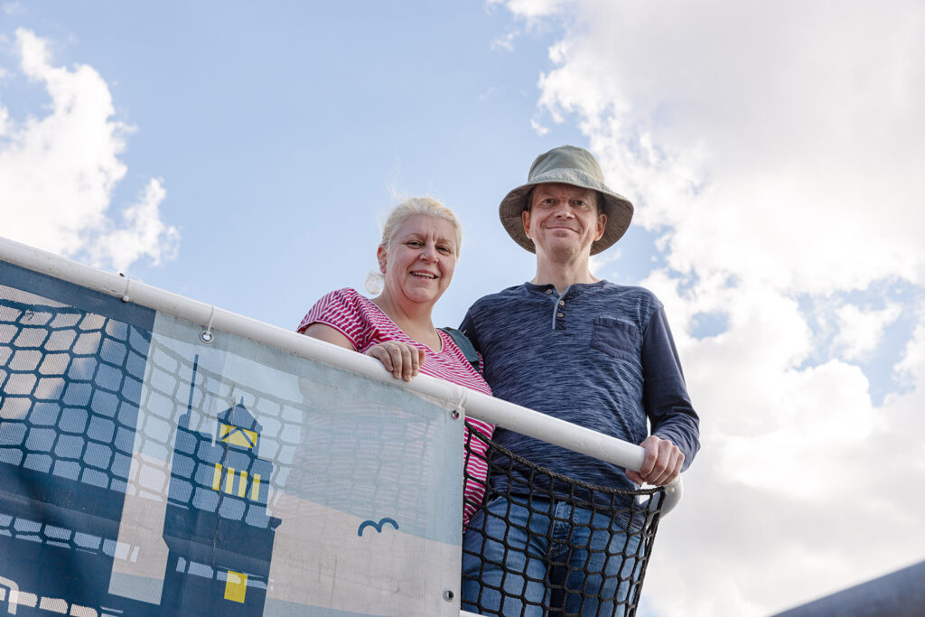 Auf einer erhöhten Plattform mit Netzgeländer stehen zwei Personen und blicken nach unten in Richtung Kamera. Eine Person trägt ein rot-weiß gestreiftes Oberteil, die andere ein blaues langärmliges Oberteil und einen breitkrempigen Hut. Unter ihnen hängt ein Banner mit einer Illustration eines Leuchtturms und eines fliegenden Vogels. Der Himmel im Hintergrund ist hell und leicht bewölkt.