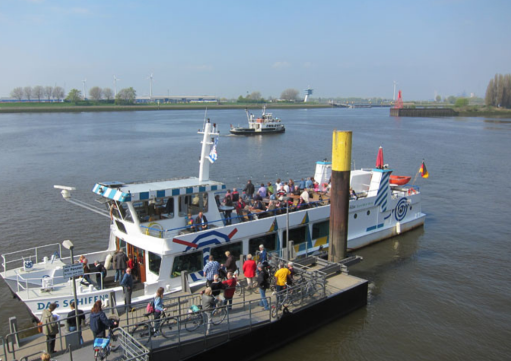 Ein Fahrgastschiff am Anleger. Fahrgäste steigen ein und aus. Im hintergrund ist ein weiteres Schiff zu sehen und der markante Radarturm am Lankenauer Höft.