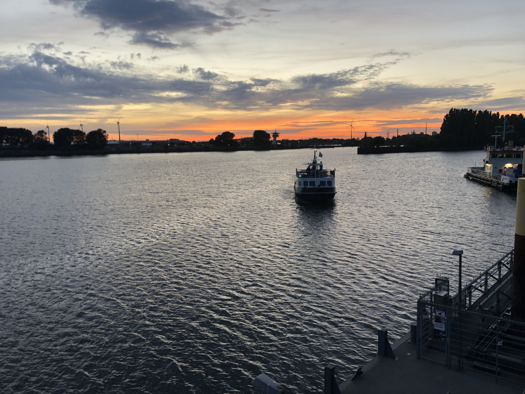 Auf einem Fluss nähert sich in Abenddämmerung ein Fahrgastschiff dem Anleger. Im Hintergrund ist das andere Flussufer im Sonnenuntergang zu erkennen.